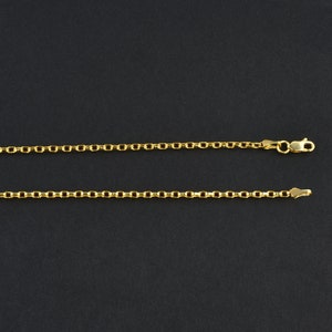 May include: Two gold-colored necklaces with a delicate chain design are displayed against a dark background. The necklaces are made of small, connected links, with one featuring a clasp. The chains are likely made of gold or a gold-colored metal.