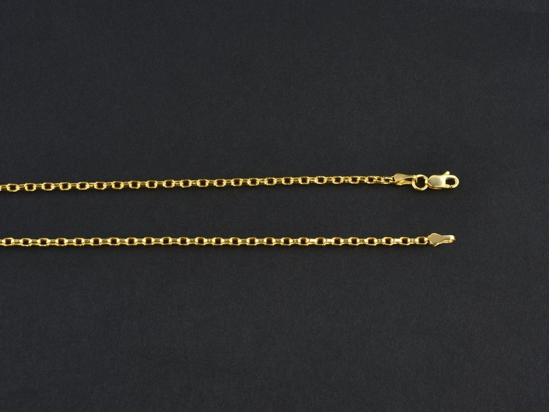 May include: Two gold-colored necklaces with a delicate chain design are displayed against a dark background. The necklaces are made of small, connected links, with one featuring a clasp. The chains are likely made of gold or a gold-colored metal.