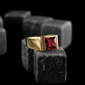 Könnte beinhalten: Ein goldener Ring mit einem quadratischen, roten Edelstein und kleinen Akzentsteinen. Der Ring liegt auf einem dunkelgrauen Stein vor schwarzem Hintergrund. Das Band des Rings hat ein schlichtes, rechteckiges Design.