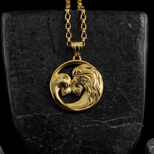 May include: Gold-colored pendant necklace featuring a lion and a serpent kissing within a circular frame. The pendant hangs from a delicate chain. The necklace is displayed on a dark, textured surface.
