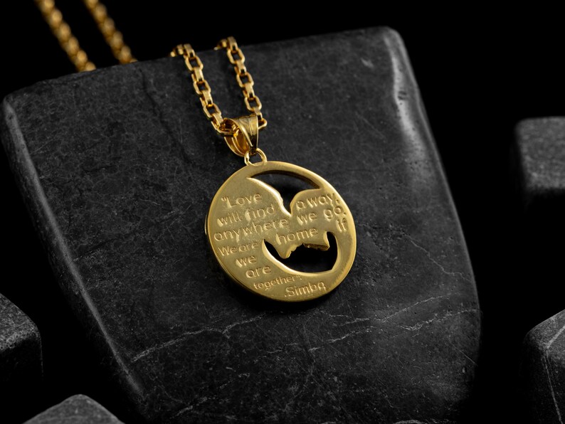 May include: A gold-colored pendant necklace with a circular charm. The charm features a cutout silhouette and engraved text that reads "Love will find a way anywhere we go if we are together Simba."