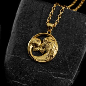 May include: A gold-colored pendant necklace featuring a circular pendant with two lion figures facing each other. The pendant hangs from a delicate gold chain. The necklace is displayed on a dark, textured surface.