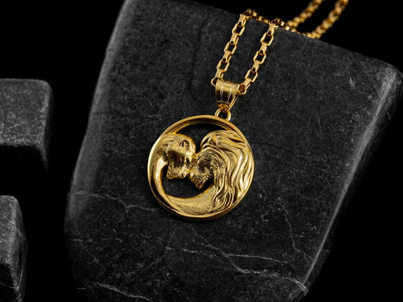 May include: A gold-colored pendant necklace featuring a circular pendant with two lion figures facing each other. The pendant hangs from a delicate gold chain. The necklace is displayed on a dark, textured surface.