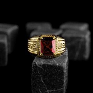 May include: A gold-colored ring with a large, rectangular, red gemstone. The ring has a decorative Greek key design on the sides and around the gemstone. The ring is displayed on a dark, textured surface against a black background.
