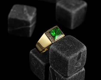 May include: A gold ring with a large, square, emerald green gemstone and smaller, clear gemstones set around the edge of the band.