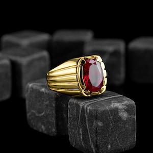 Puede incluir: Un anillo de oro con una piedra preciosa roja grande de forma ovalada. El anillo está colocado sobre una superficie negra texturizada.