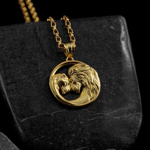 May include: Gold-colored pendant necklace featuring a circular design with two kissing figures. The pendant hangs from a delicate gold chain. The necklace is displayed on a dark, textured surface, creating a striking contrast.