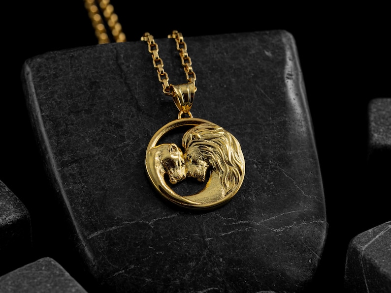 May include: Gold-colored pendant necklace featuring a circular design with two kissing figures. The pendant hangs from a delicate gold chain. The necklace is displayed on a dark, textured surface, creating a striking contrast.