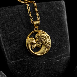 May include: Gold-colored pendant necklace featuring a lion and a human face kissing within a circular frame. The pendant hangs from a delicate chain. The necklace is displayed on a dark, textured surface.