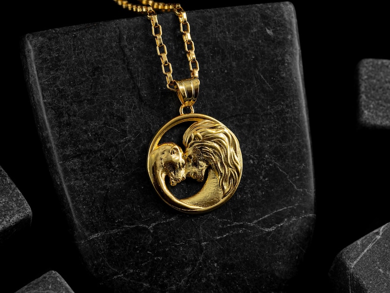 May include: Gold-colored pendant necklace featuring a lion and a human face kissing within a circular frame. The pendant hangs from a delicate chain. The necklace is displayed on a dark, textured surface.