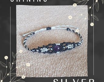 Puede incluir: Pulsera de macramé con una gema púrpura central, un patrón tejido en blanco y negro y cuentas doradas. El texto "SHINING SILVER" se muestra en letras blancas sobre un fondo negro.