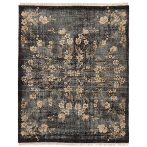 May include: A rectangular, dark gray rug with a floral pattern in shades of beige and cream. The rug has a distressed look, with a fringe along the edges. The floral design is symmetrical, with flowers and leaves arranged in a central medallion.