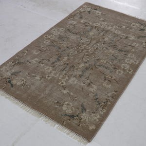 May include: Rectangular, beige rug with a floral pattern. The rug features a central floral design with surrounding floral and leaf motifs in shades of beige, brown, and blue-green. The rug has a fringe on the short ends.