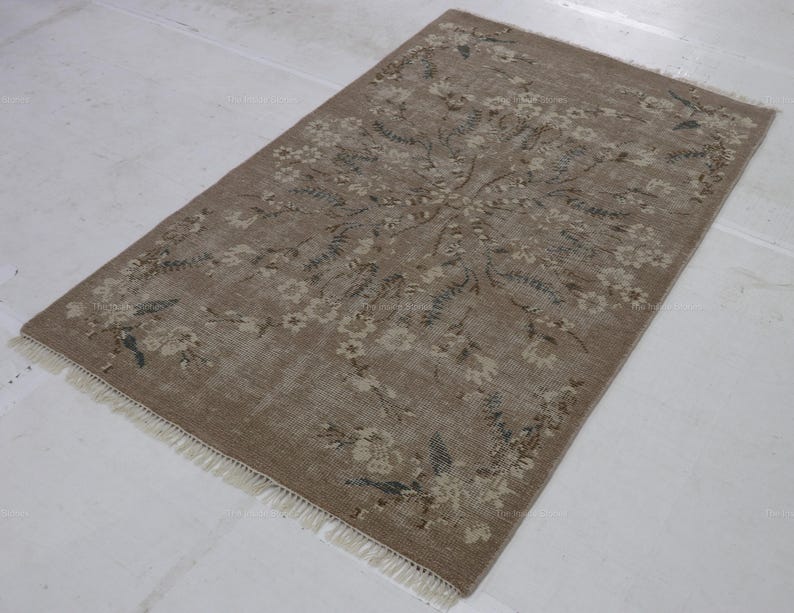 May include: Rectangular, beige rug with a floral pattern. The rug features a central floral design with surrounding floral and leaf motifs in shades of beige, brown, and blue-green. The rug has a fringe on the short ends.
