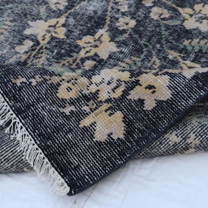 May include: A section of a distressed, floral-patterned rug. The rug features a dark blue background with faded beige and green floral accents. The edge of the rug is folded over, revealing a white fringe.