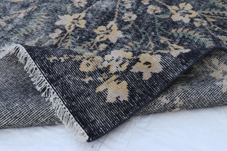 May include: A section of a distressed, floral-patterned rug. The rug features a dark blue background with faded beige and green floral accents. The edge of the rug is folded over, revealing a white fringe.
