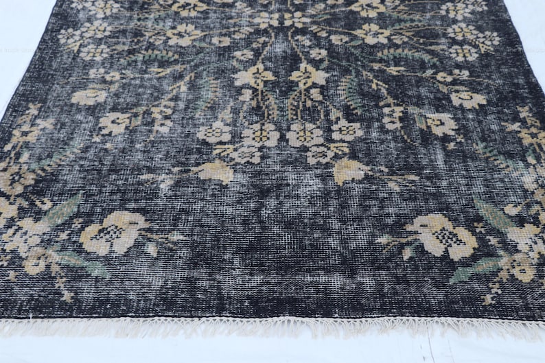 May include: A distressed black rug with a floral pattern in cream and green. The rug has a faded, vintage look with a fringed edge. The floral design is spread across the rug.
