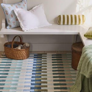 May include: A rectangular area rug featuring a repeating geometric pattern of vertical bars in shades of blue, sage green, and cream. The rug is positioned on the floor in front of a white bench with various pillows and a woven basket.