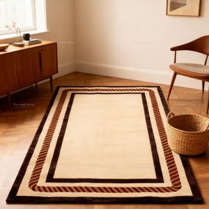 May include: Rectangular beige rug with a dark brown border and a rope-like design in shades of brown and tan. The rug is placed on a wooden floor in a room with a window and a wooden cabinet.