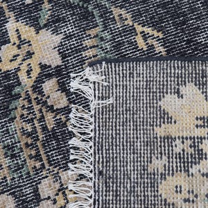 May include: Close-up of a distressed, patterned rug with a black and white background. The rug features a floral design in shades of beige and green. The edge of the rug has a fringe.
