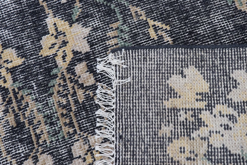 May include: Close-up of a distressed, patterned rug with a black and white background. The rug features a floral design in shades of beige and green. The edge of the rug has a fringe.