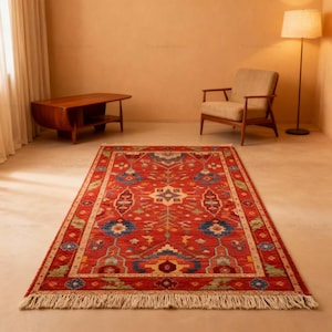 May include: A rectangular, red area rug with an intricate geometric pattern in blue, green, and yellow. The rug has a cream-colored border and fringe along the edges. The rug is placed on a light-colored floor.