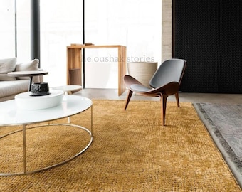 May include: A large, rectangular, mustard-yellow area rug with a textured, grid-like woven pattern. The rug features a dense, looped pile construction and covers the floor space in front of a white coffee table and a dark-toned lounge chair.