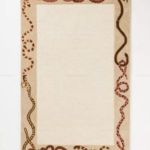 May include: A rectangular area rug featuring a solid cream-coloured centre panel framed by a wide beige border. The border is decorated with a continuous pattern of winding snakes in various shades of brown, tan, and pink, creating a detailed motif.