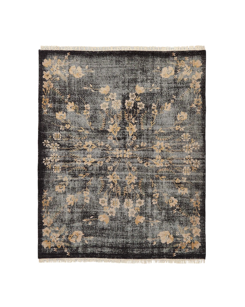 May include: A rectangular, dark gray rug with a floral pattern in shades of beige and cream. The rug has a distressed look, with a fringe along the edges. The floral design is symmetrical, with flowers and leaves arranged in a central medallion.