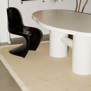 May include: A glossy black cantilever chair with a curved silhouette positioned next to a large white oval dining table featuring two thick cylindrical pedestal legs and a horizontal support beam, all set upon a beige area rug with rounded corners.