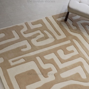 May include: Beige and cream-colored rectangular rug with a geometric pattern. The rug features a repeating design of interconnected shapes in a lighter cream color against a beige background. A white upholstered bench with dark brown legs is in the background.