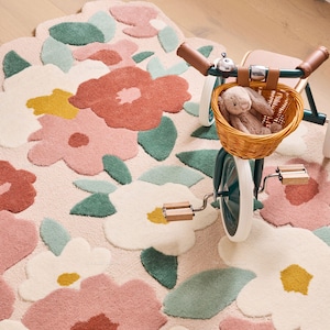 Könnte beinhalten: Ein floraler Teppich mit gewelltem Rand zeigt große, farbenfrohe Blumen- und Blattmuster in Rosa-, Weiß-, Grün- und Gelbtönen. Ein kleines Dreirad mit einem Weidenkorb und einem Stoffhasen befindet sich auf dem Teppich.