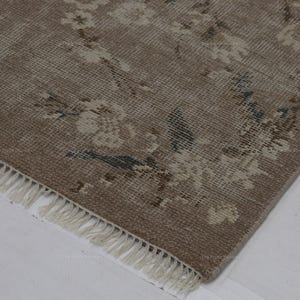 May include: A corner view of a distressed, vintage rug with a floral pattern. The rug features a muted color palette of beige, cream, and touches of blue. The edge has a fringe detail.
