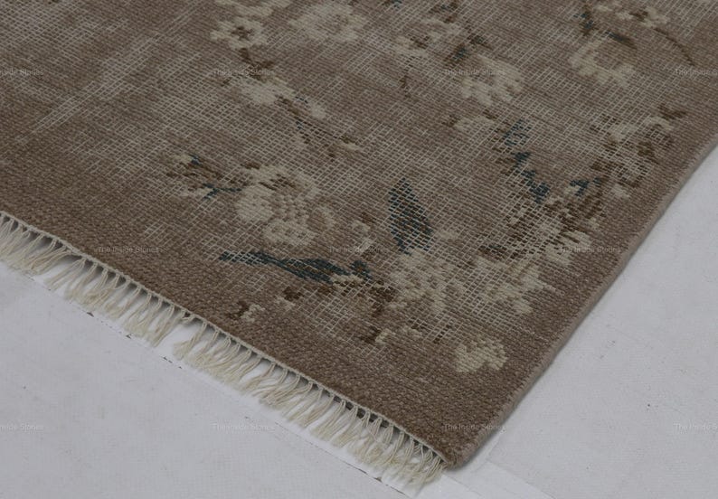 May include: A corner view of a distressed, vintage rug with a floral pattern. The rug features a muted color palette of beige, cream, and touches of blue. The edge has a fringe detail.