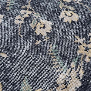 May include: Close-up of a distressed, vintage rug with a dark gray background and a floral pattern in beige and green. The rug has a woven texture and a faded appearance, suggesting age and use.