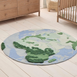 May include: A circular area rug featuring a tufted map of the Earth. The design displays continents in varying shades of green and deep forest green, contrasted against oceans rendered in light blue and soft grey tones with a textured surface.