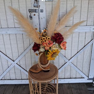 May include: A tan leather cowboy boot serves as a vase for a floral arrangement featuring tall beige pampas grass, deep red dahlias, bright yellow sunflowers, and orange blossoms, accented with a coiled piece of light-colored rope around the boot.