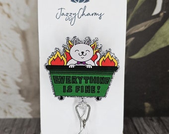 May include: A green and silver glitter badge reel with a white cat in a dumpster on fire. The cat is wearing a bow tie and the dumpster says "Everything is fine!"