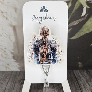 May include: A white card with the text "Jazzy Charms" and a floral design featuring a woman with long brown hair wearing a blue and white dress and armor. The card is attached to a silver retractable badge reel.