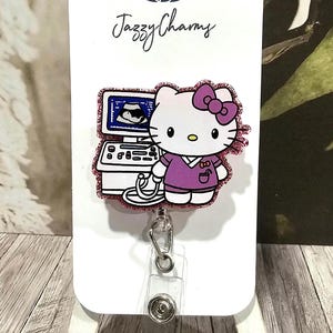 May include: Pink glitter Hello Kitty badge reel with a pink ultrasound machine and a stethoscope. The badge reel is attached to a white card with the text "Jazzy Charms".