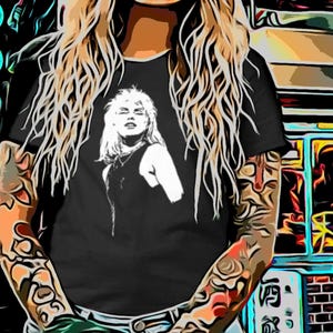 Puede incluir: Camiseta negra con un gráfico blanco de una mujer. La persona de la imagen tiene el pelo largo y rubio y tatuajes en los brazos. El fondo incluye texto japonés y escaparates coloridos.