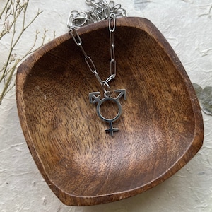Op de afbeelding: Een zilveren ketting met een transgender symbool hanger. De ketting wordt getoond in een houten kom op een wit, getextureerd oppervlak.