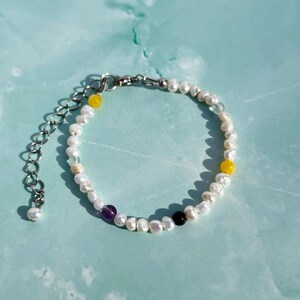 May include: A bracelet featuring small white irregular freshwater pearls interspersed with single yellow, purple, and black beads. The piece includes a silver-toned metal chain extender with a lobster clasp and a small pearl at the end.