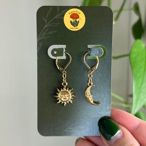 Puede incluir: Pendientes dorados con dise&ntilde;os de sol y luna. El pendiente de sol tiene una cara detallada con rayos, mientras que el pendiente de luna representa una luna creciente con una cara. Los pendientes se muestran en una tarjeta verde oscuro con una pegatina "Earth Star".