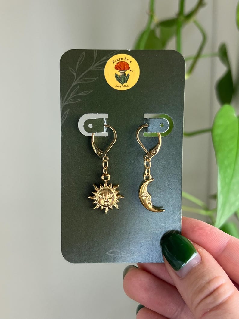 Puede incluir: Pendientes dorados con dise&ntilde;os de sol y luna. El pendiente de sol tiene una cara detallada con rayos, mientras que el pendiente de luna representa una luna creciente con una cara. Los pendientes se muestran en una tarjeta verde oscuro con una pegatina "Earth Star".