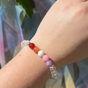 Peut inclure: Un bracelet avec des perles blanches, rouges, orange, blanches, roses, violettes et blanches. Le bracelet est au poignet d'une personne.