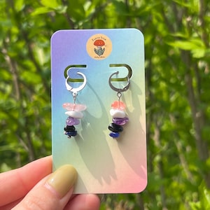 Op de afbeelding: Een paar bungelende oorbellen met zilverkleurige hoepels. Elke oorbel heeft een stapel kleurrijke edelstenen in roze, paars, zwart en blauw. De oorbellen worden gepresenteerd op een pastelkleurige kaart met de tekst "Earth Star".