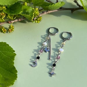 May include: A pair of silver-toned hoop earrings featuring dangling chains with small white pearls, translucent pink beads, and black beads. One earring includes a silver crescent moon charm, while the other features a four-pointed star charm.