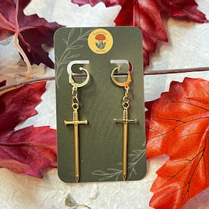 Puede incluir: Pendientes dorados con forma de espada colgados de una tarjeta verde con un logotipo. Los pendientes tienen un cierre de palanca. El fondo incluye hojas rojas y naranjas.