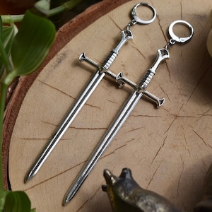 May include: A pair of silver sword earrings with a detailed design. The earrings are hanging from a wooden surface.